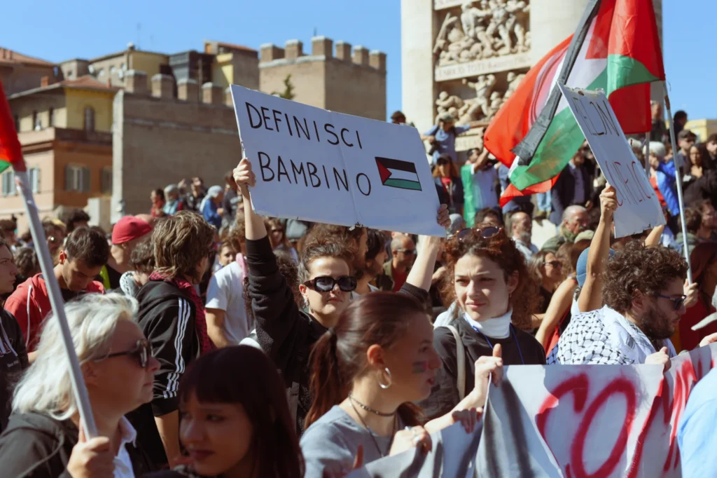 Una manifestante solleva un cartello con la scritta "Definisci bambino" con una bandiera palestinese disegnata, in mezzo a una folla di persone che partecipano a una manifestazione in piazza. Sullo sfondo sono visibili bandiere palestinesi e un monumento. Foto di Francesco Bignardi.