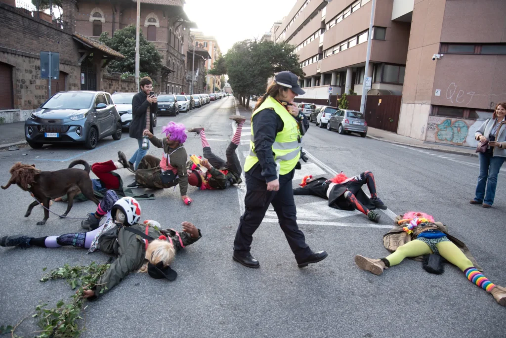 Un gruppo di attivisti in costumi colorati e stravaganti è sdraiato sull'asfalto di un incrocio in un'azione di protesta. Una vigilessa in gilet giallo attraversa la scena. Un cane si aggira tra i manifestanti. Foto di Francesco Bignardi.