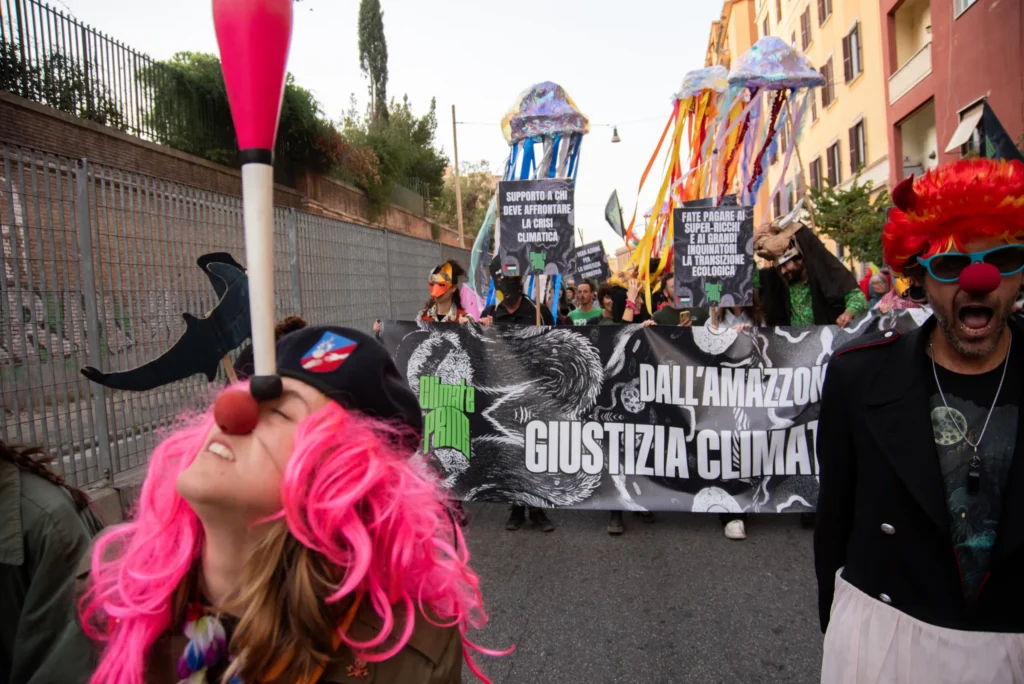 Corteo per la giustizia climatica: in primo piano una persona con parrucca rosa e naso da clown equilibra un birillo sul mento. Sullo sfondo uno striscione recita "Dall'Amazzo[nia] Giustizia Clima[tica]", con grandi sculture di meduse colorate portate in corteo. Foto di Francesco Bignardi.