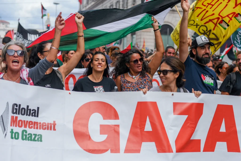 In primo piano un gruppo di manifestanti sorridenti e a pugno alzato porta uno striscione bianco con la scritta "Global Movement to Gaza" e la parola "GAZA" in grandi lettere rosse. Sopra le loro teste sventola una bandiera palestinese. Sullo sfondo una folla densa con altre bandiere. Foto di Francesco Bignardi.